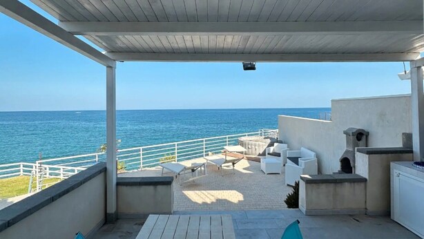 A covered outdoor area with a view of the sea, featuring a table, chairs, and a barbecue grill.