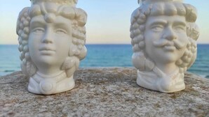 Deux bustes en céramique blanche, ornés de coiffures complexes, sont posés sur une surface en pierre, avec en arrière-plan une vue dégagée sur l’océan.