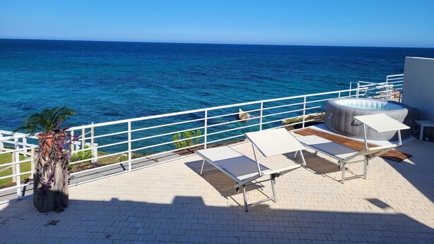Un balcon avec vue sur l’océan, des fauteuils de jardin blancs et une petite table.
