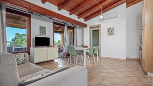 A living area with a sofa, TV, dining table, chairs, and a balcony with a view. - Holiday Home 'Orizzonti 33' with Sea View, Private Terrace and Air Conditioning (Golfo Aranci)