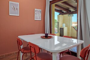 Un coin repas avec une table blanche et des chaises rouges, des tableaux encadrés au mur et une vue à travers une porte vitrée.