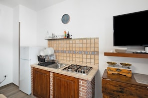 Una cocina compacta con estufa a gas, un lavamanos, un refrigerador, un reloj y un televisor sobre un pedestal.