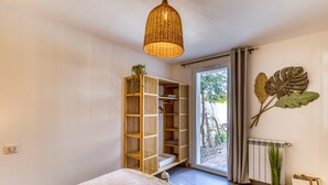 Een slaapkamer met een bed, een rieten plafondlamp, een houten boekenplank, een grote plant en uitzicht door een glazen deur.
