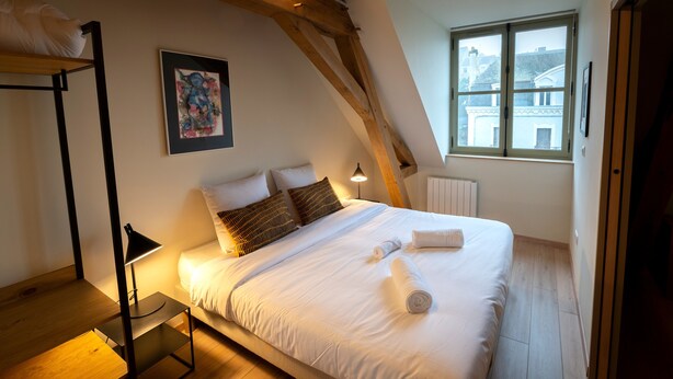 Une chambre à coucher avec un lit, des oreillers, une table de chevet, une lampe, un radiateur et une fenêtre.