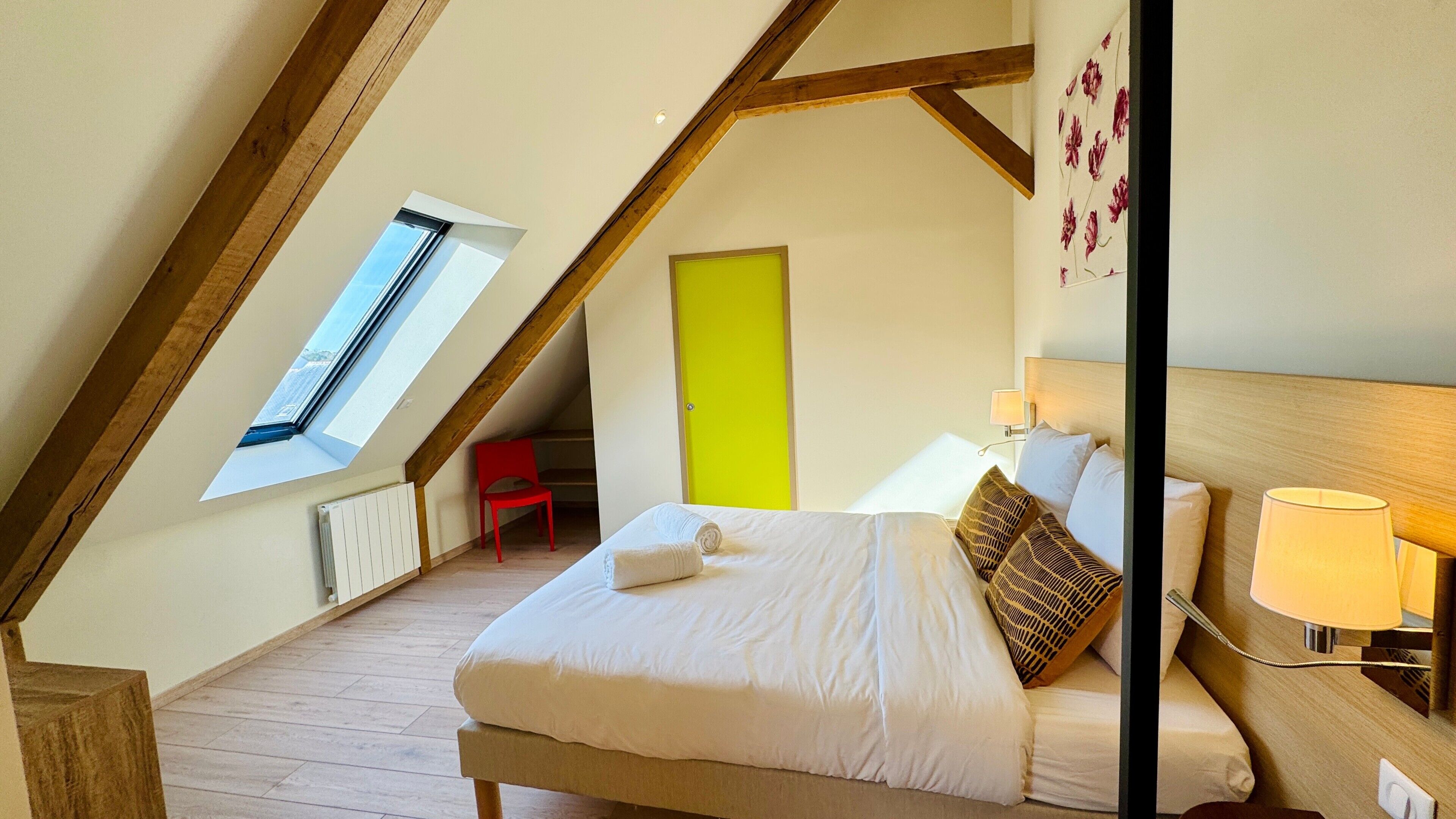 A bedroom with a sloped ceiling, a bed with white bedding, a red chair, a bright green door, and a window with a view.