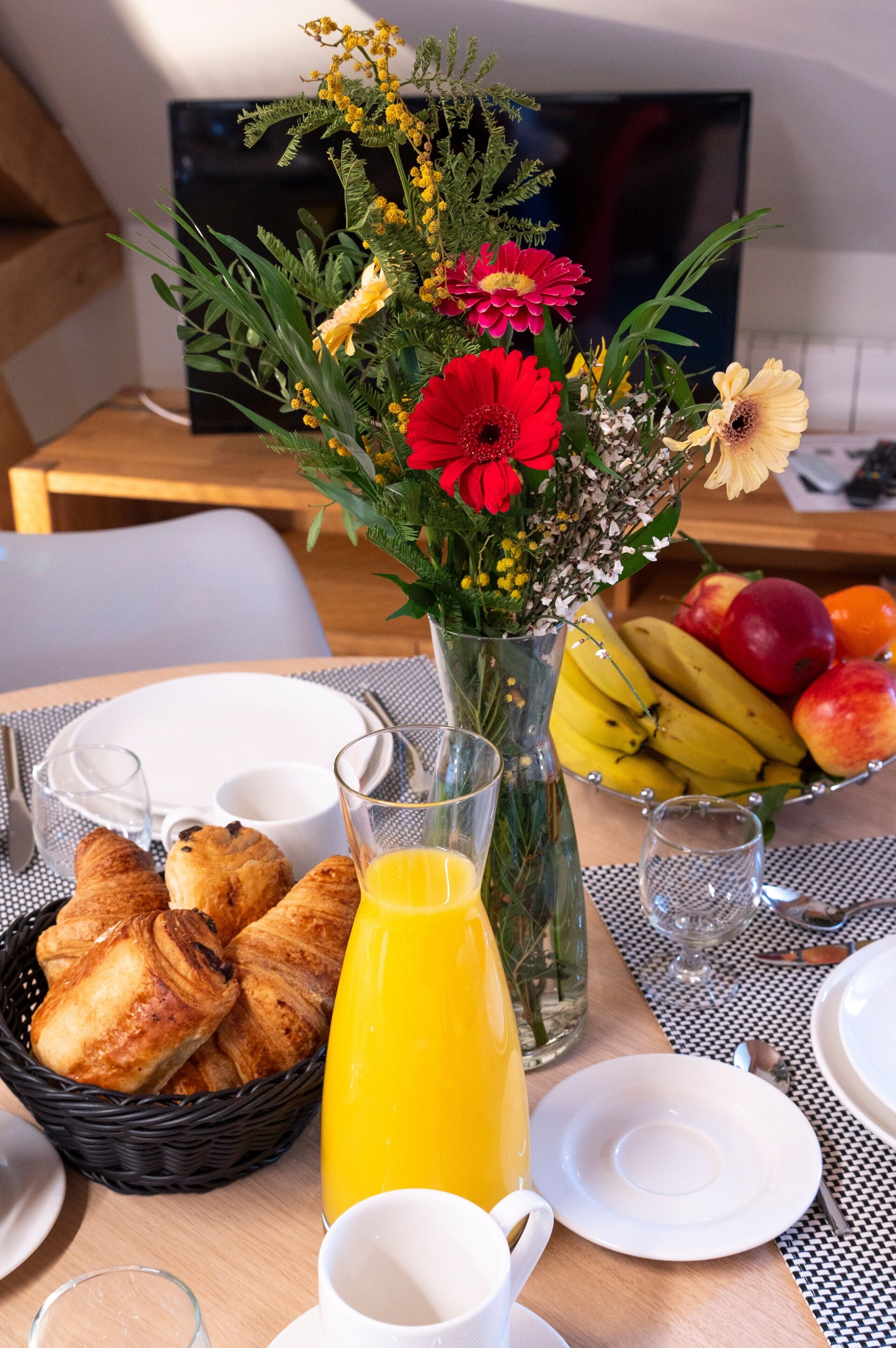 A breakfast table with croissants, a glass of orange juice, a fruit bowl, and a vase with flowers.