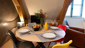 A dining area with a round table, chairs, a vase with flowers, a basket of bread, and a jug of orange juice.