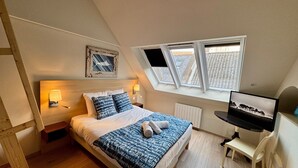 Quarto com teto inclinado, cama com roupa de cama estampada, duas luminárias fixadas na parede, um quadro emoldurado e uma TV sobre um suporte.