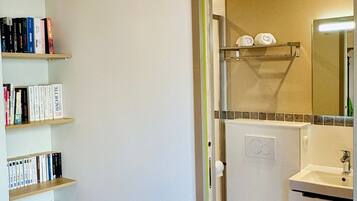 A bathroom with a toilet, sink, and mirror. There is a bookshelf with books to the left.