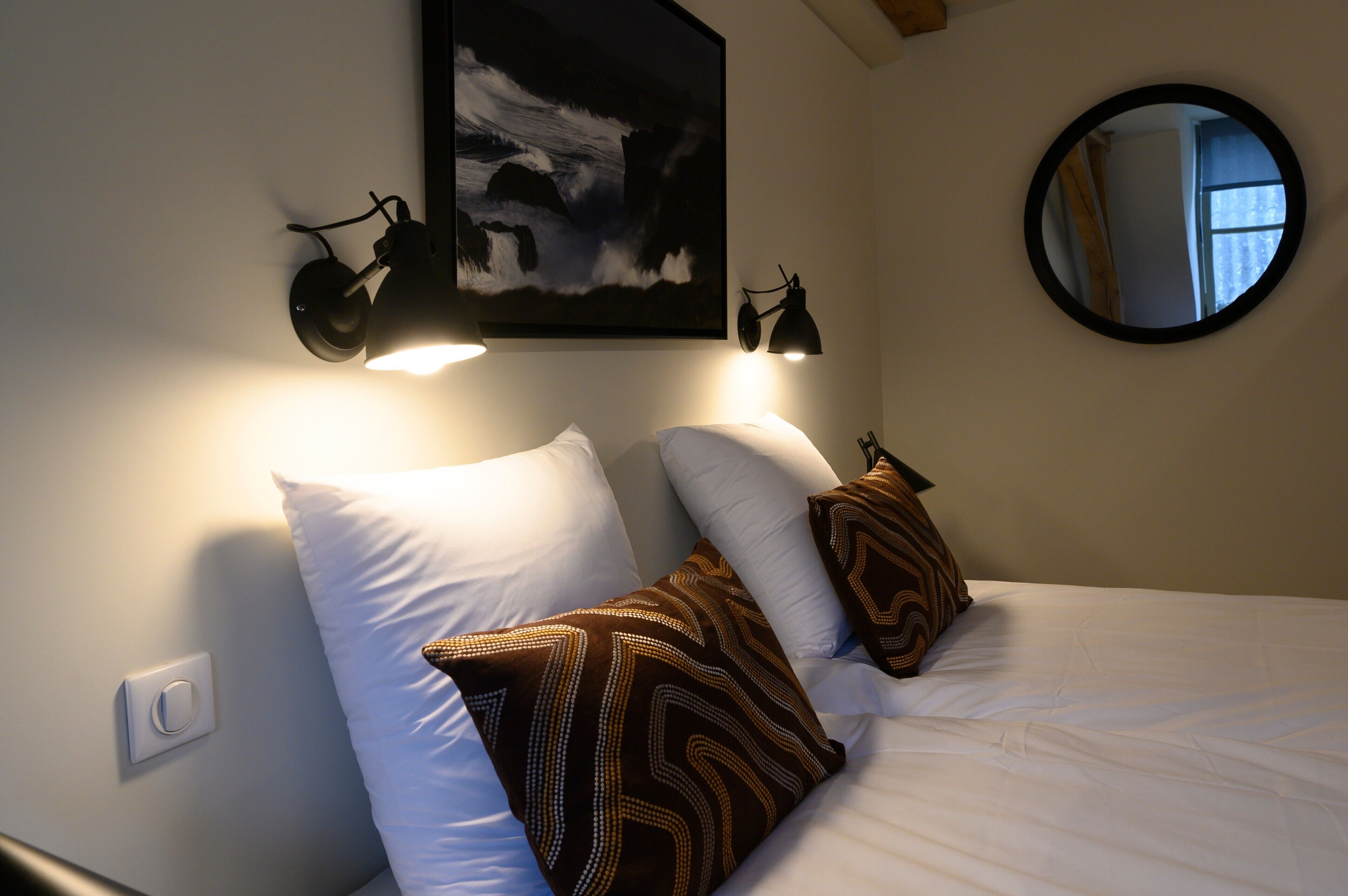 A bed with white and patterned pillows, a wall-mounted lamp, and a framed picture above the bed.