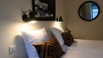 A bed with white and patterned pillows, a wall-mounted lamp, and a framed picture above the bed.