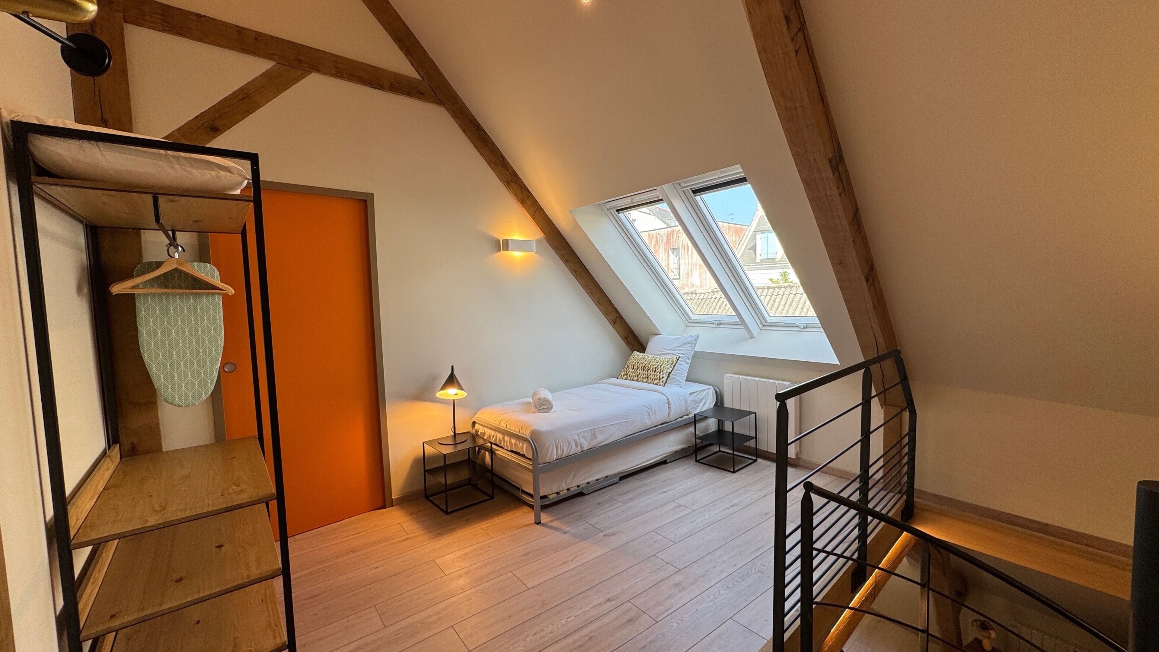 Et soveværelse under skrå loft med seng, et lille bord, en knagerække og et vindue med udsigt til bygninger.