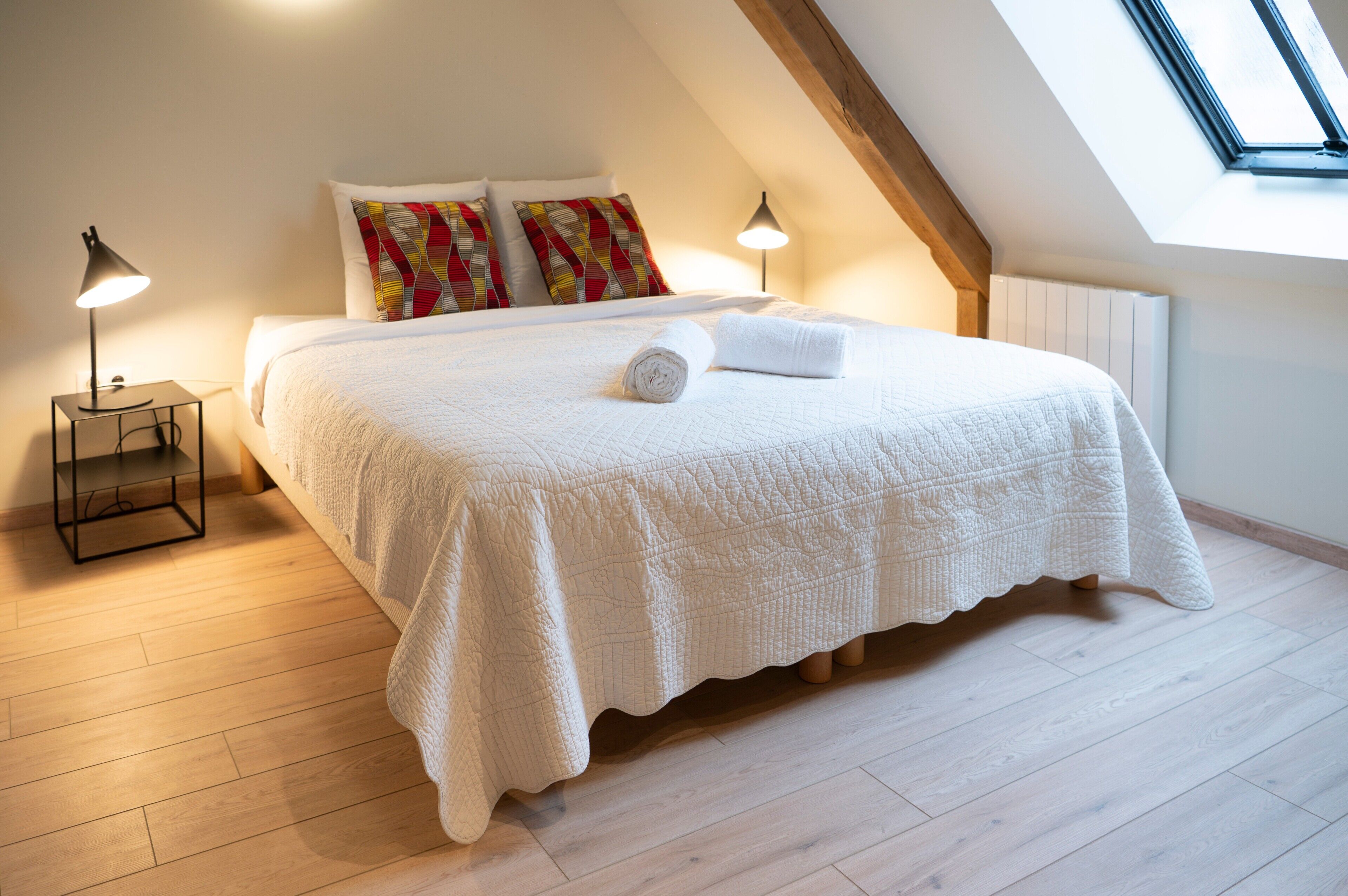 Quarto com teto inclinado, uma cama com colcha branca, duas luminárias de apoio e um domo de vidro.