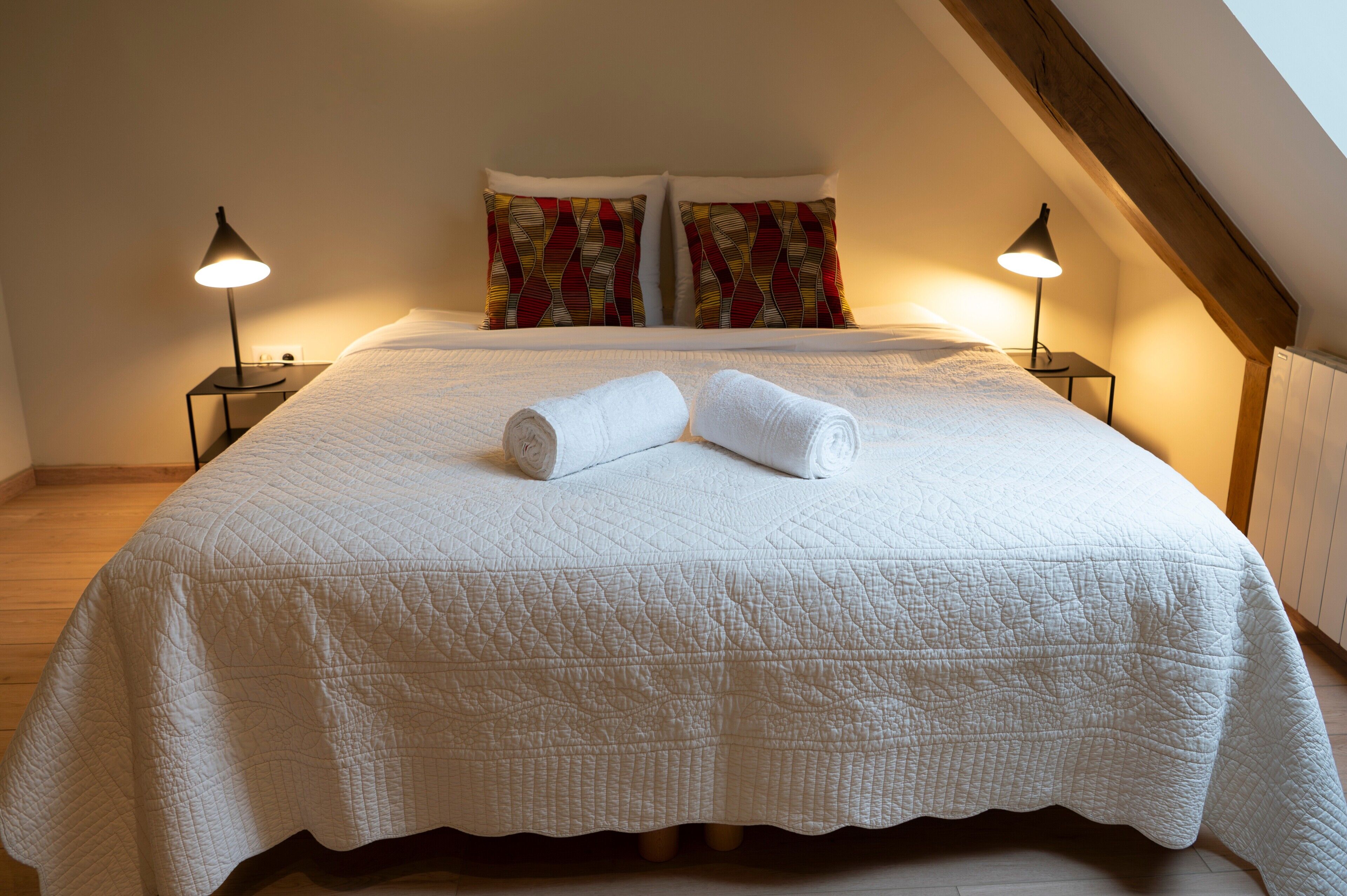 Ein ordentlich bezogenes Bett mit einer weißen Tagesdecke, zwei Kissen mit geometrischem Muster und zwei Nachttischlampen.