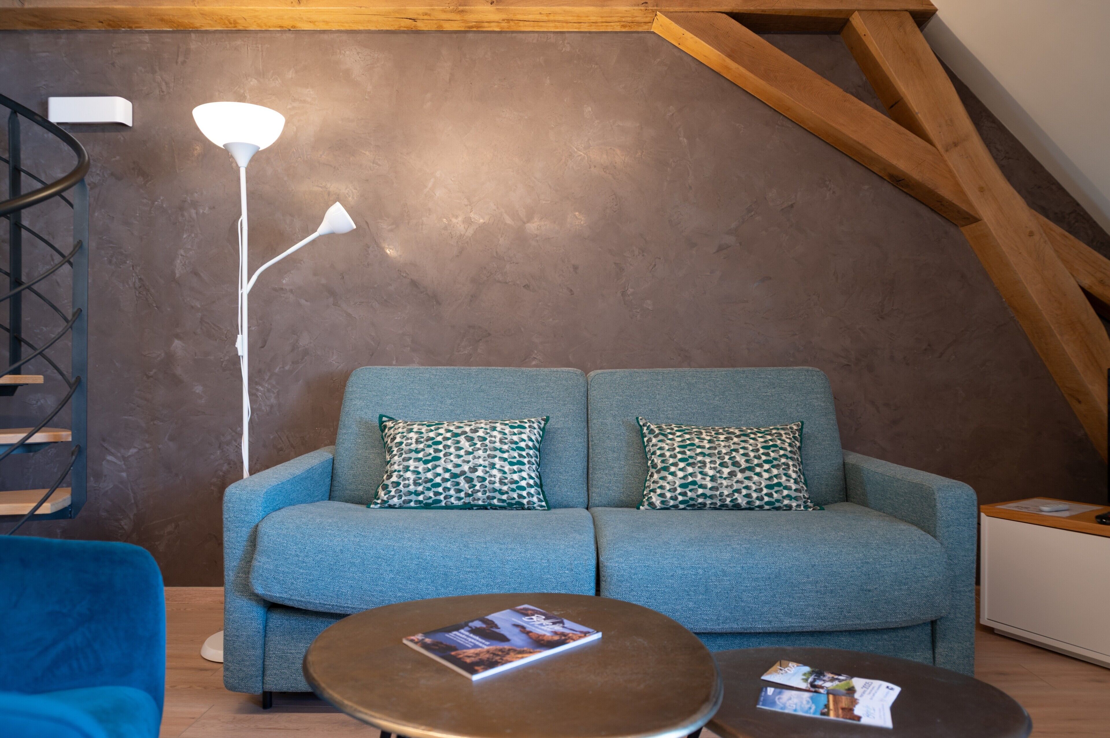 A modern loft-style room with a blue sofa, patterned pillows, a round coffee table, a floor lamp, and a wooden ceiling.
