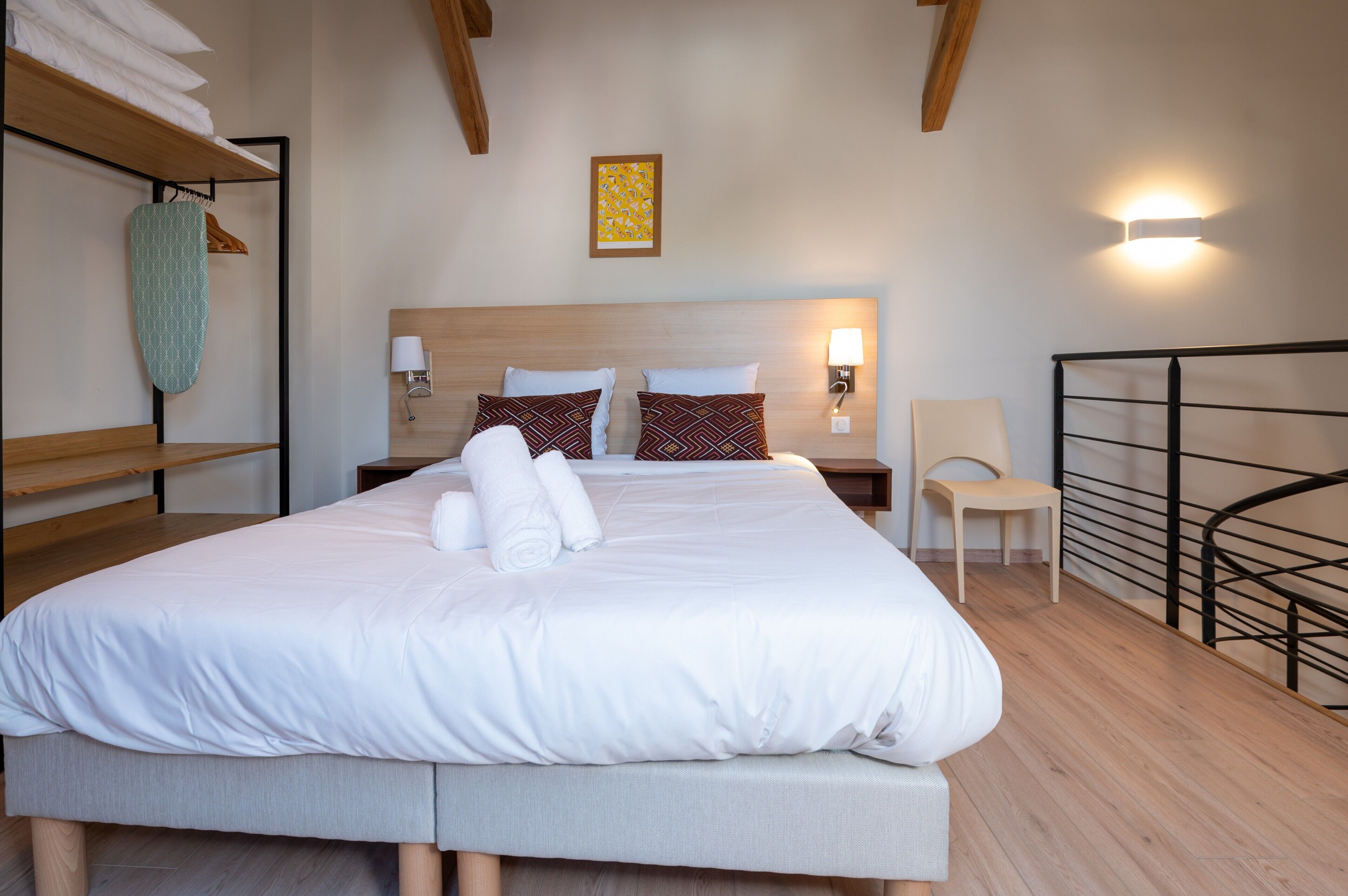 Een netjes opgemaakt bed met kussens, een bedlampje, een stoel en een wanddecoratie.