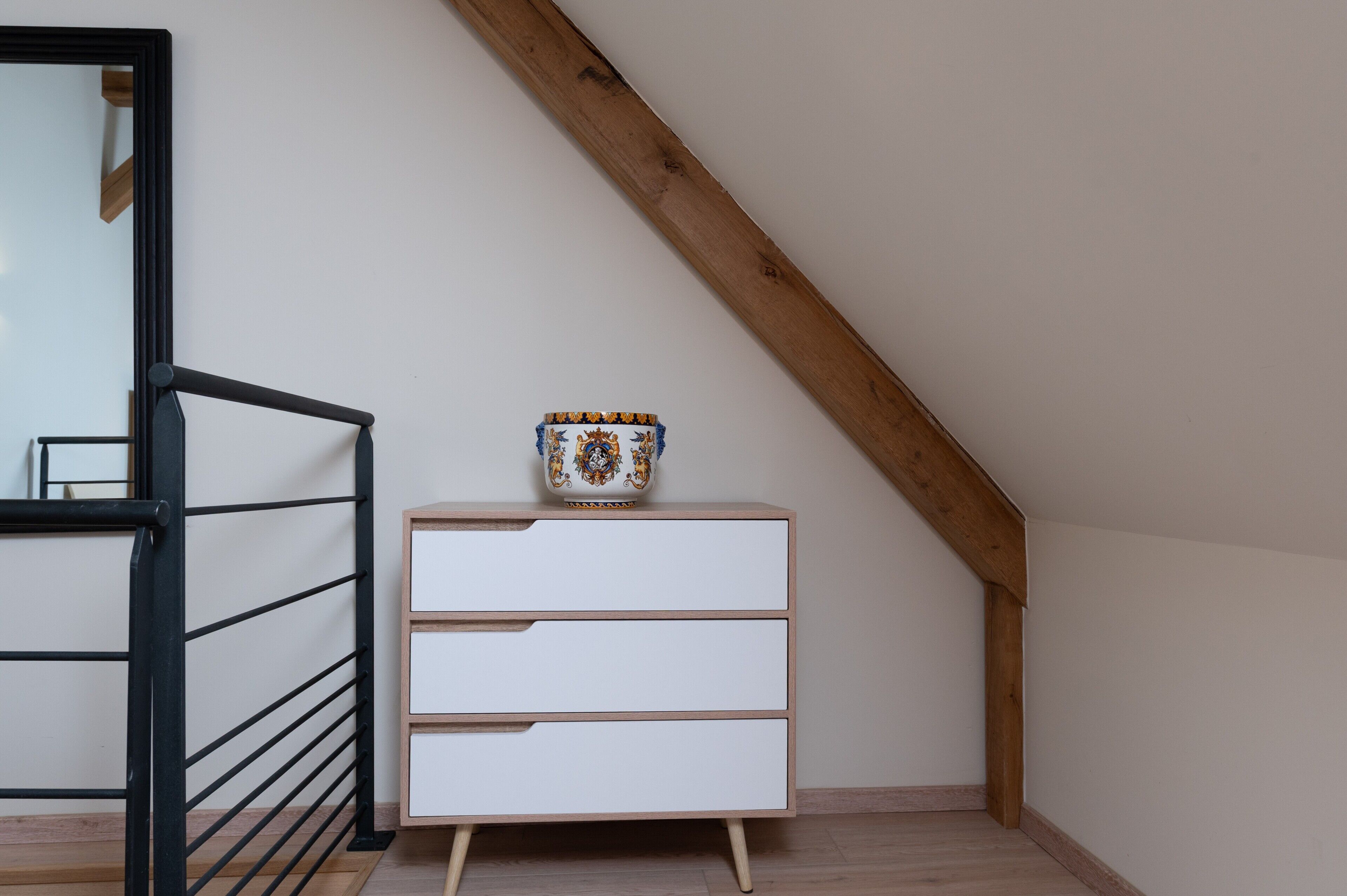 Een kamer met een witte ladekast, een decoratieve vaas, een zwarte metalen balustrade en een houten plafondbalk.