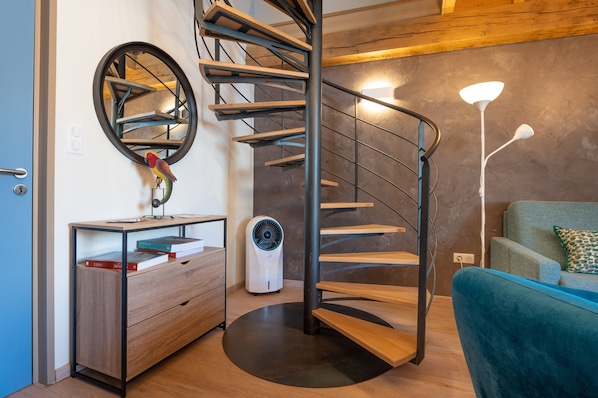 A modern interior with a spiral staircase, a round mirror, a desk with books, a standing fan, and a blue sofa.
