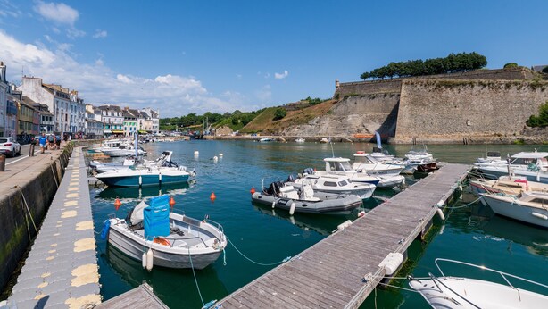 A harbor with boats docked at a wooden pier, a stone wall, and buildings along the waterfront.