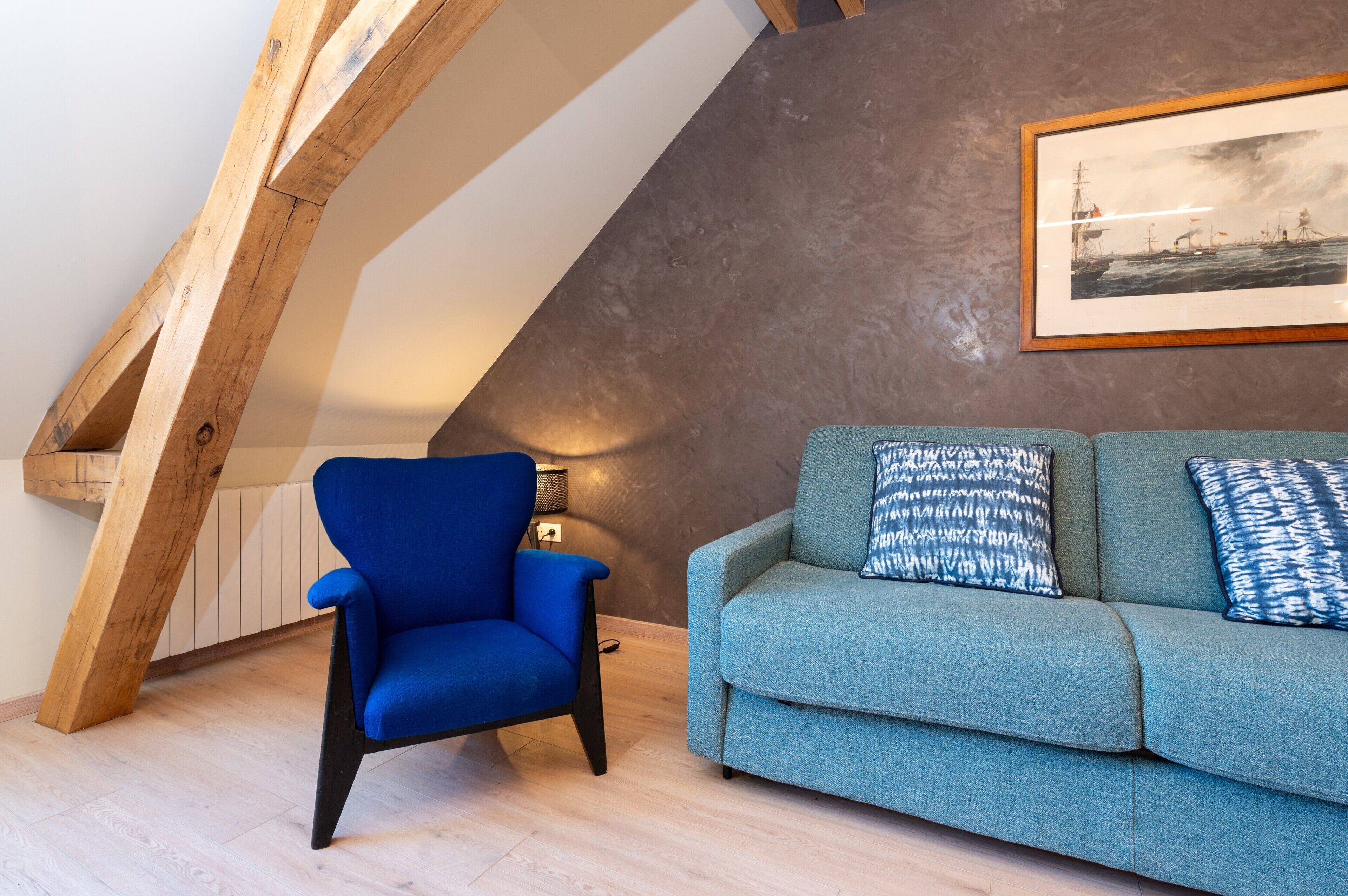 Ein Zimmer mit geneigter Decke, einem blauen Sessel, einem blaugrünen Sofa mit gemusterten Kissen und einem gerahmten Bild an der Wand.