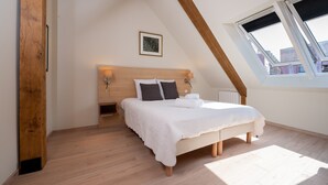 A bedroom with a sloped ceiling, a bed with a white bedspread, two wall-mounted lamps, a framed picture, and a window with blinds.