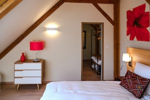 A bedroom with a sloped ceiling, a bed with white bedding, a red lamp, a white dresser, and a large red flower painting on the wall. - Seigneurie Fouquet - Miscou Apartment with Wi-Fi (Le Palais)