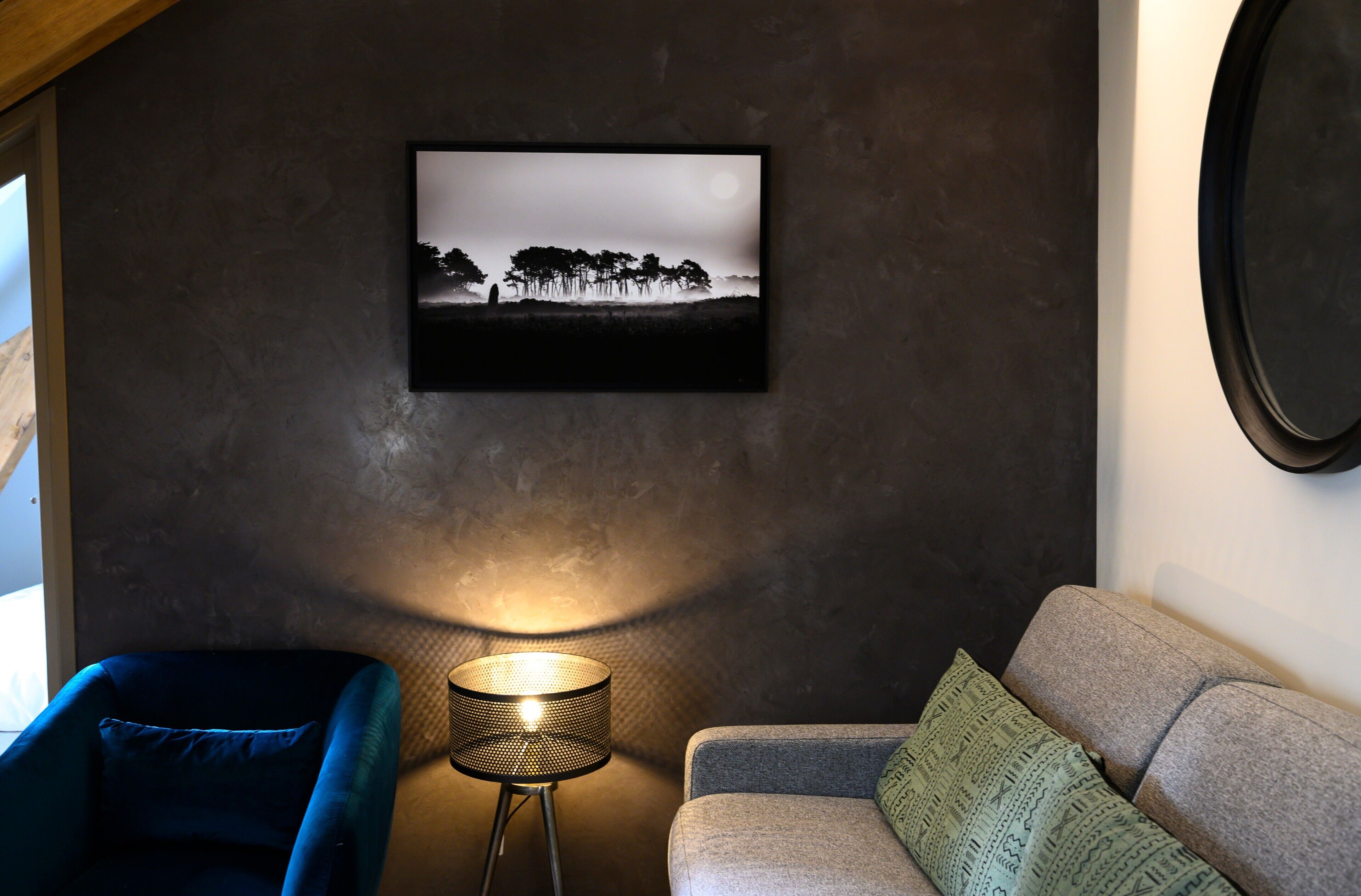 Ein Zimmer mit dunkler Wandstruktur, einem gerahmten Schwarz-Weiß-Landschaftsfoto, einer runden Stehlampe, einem blauen Sessel und einem grauen Sofa mit gemusterten Kissen.