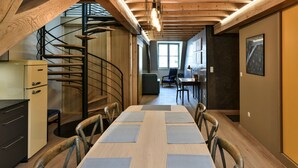 A dining area with a long table, chairs, a spiral staircase, and a kitchen area.