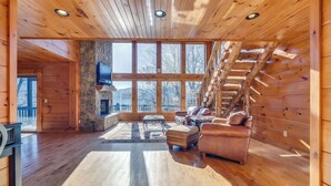 A spacious cabin interior with a stone fireplace, wooden walls, and a large window overlooking a snowy landscape. - Lakefront Mountain Retreat on Watauga Lake (Butler)