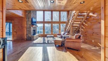 A spacious cabin interior with a stone fireplace, wooden walls, and a large window overlooking a snowy landscape.
