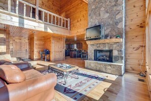 A spacious wooden cabin with a stone fireplace, a television mounted above it, and a dining area in the background.
