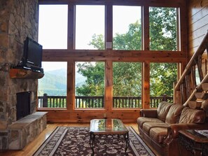 Ein Blockhaus-Interieur mit Kamin aus Stein, einem Ledersofa, einem Glastisch und großen Fenstern mit Blick auf die Berge.