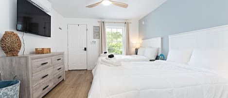 Habitación de hotel con piso de madera, ventilador de techo, televisor montado en la pared, cómoda de madera, cama con ropa blanca y ventana con cortinas.