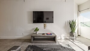 Ein modernes Wohnzimmer mit einem flachbildfernseher an der Wand, einem kleinen Konsolentisch mit Dekorationsobjekten, einem großen Teppich und einem Blumentopf am Fenster.