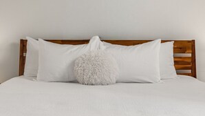 Ein Bett mit Holz Kopfteil, weißer Bettwäsche und einem flauschigen weißen Kopfkissen.