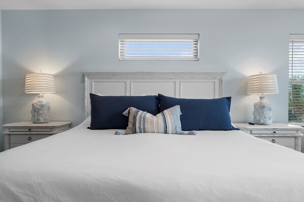 A neatly made bed with two pillows, a bedside table with a lamp, and a window with blinds.