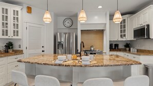 Une cuisine moderne avec des meubles blancs, un îlot en granit doté de tabourets, des appareils en acier inoxydable et des luminaires suspendus.