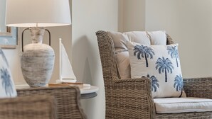 Une chaise en osier avec un coussin orné de motifs de palmiers, un vase blanc et une lampe posée sur une petite table d’appoint.