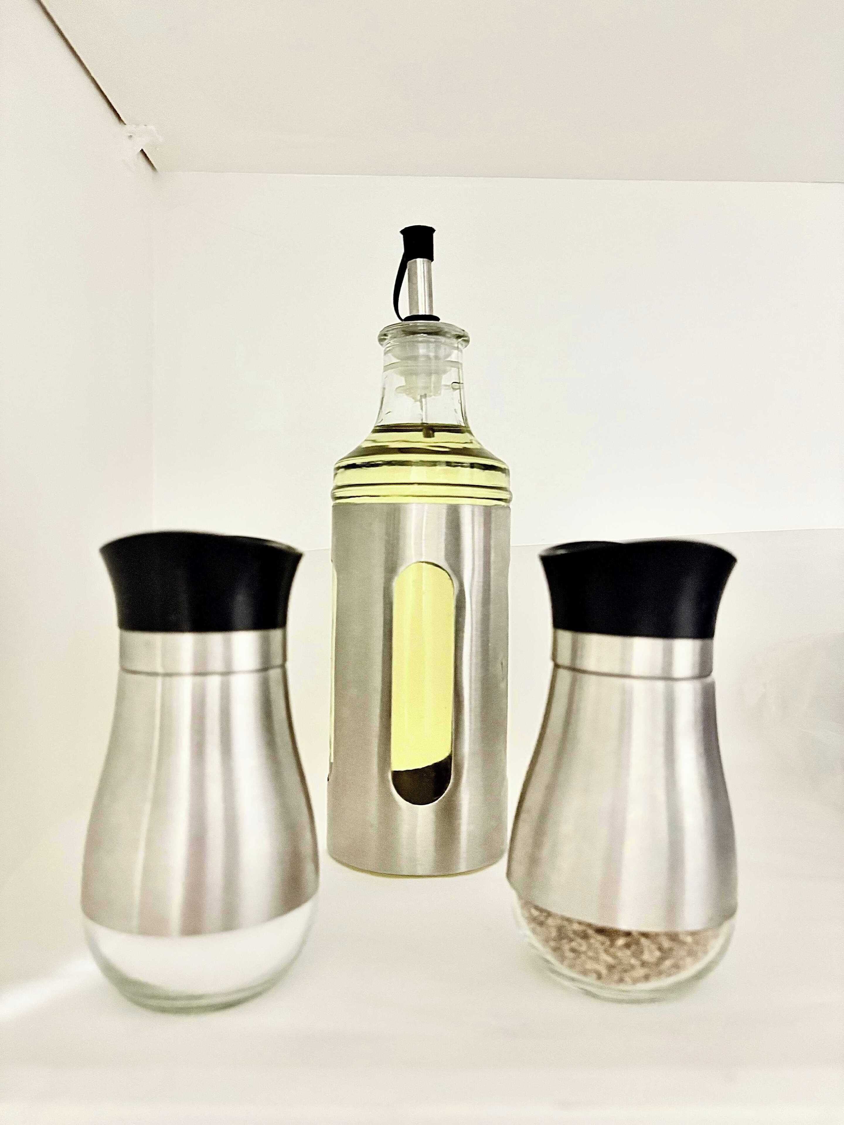 Tres recipientes de especias con tapas negras y cuerpos metálicos, uno con una sección transparente y tapa negra, sobre una superficie blanca.