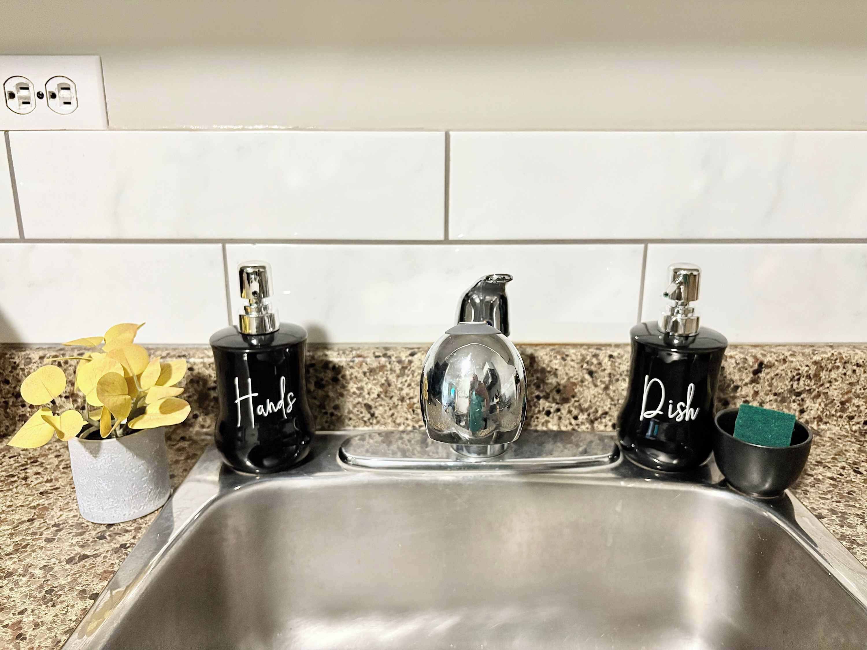A kitchen sink with two soap dispensers labeled "Hands" and "Dish" and a small plant with yellow flowers.