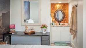 Une chambre à coucher avec un lit, une table de chevet sur laquelle se trouve un livre et un vase de fleurs, une commode avec un miroir, et une salle de bain équipée d’un lavabo et d’un porte-serviettes.