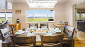 A dining area with a table set for four, surrounded by wooden chairs, a chandelier, and a view of a green landscape through a large window. - New! Home on one acre, dog friendly, private hot tub (Kalispell)