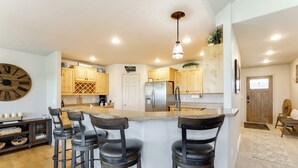 A modern kitchen with wooden cabinets, a stainless steel refrigerator, and a bar area with stools. - New! Home on one acre, dog friendly, private hot tub (Kalispell)