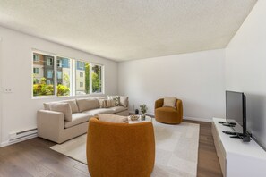 Un salon moderne avec un canapé beige, un fauteuil rond orange, un téléviseur à écran plat et une grande fenêtre donnant sur des immeubles et des espaces verts.