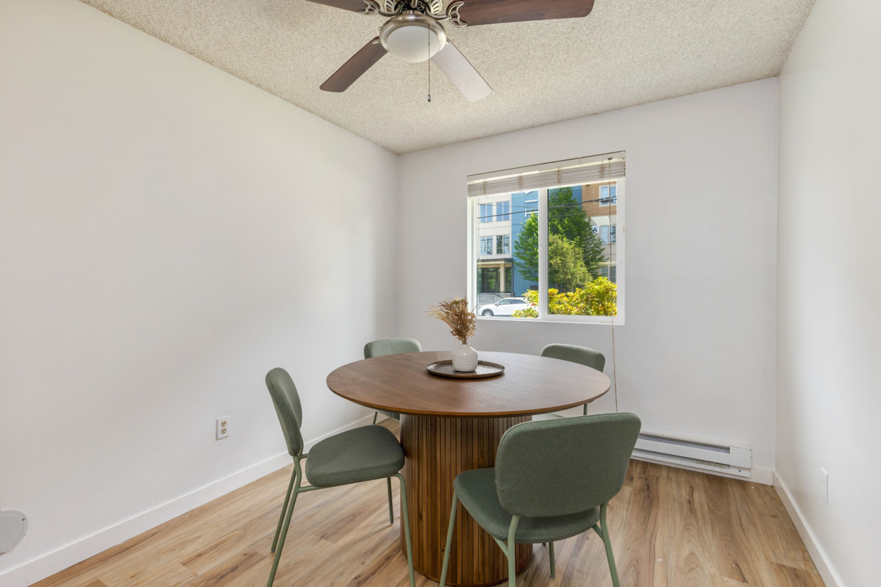 A modern dining area with a round table, four chairs, a ceiling fan, and a window with a view of buildings and trees.