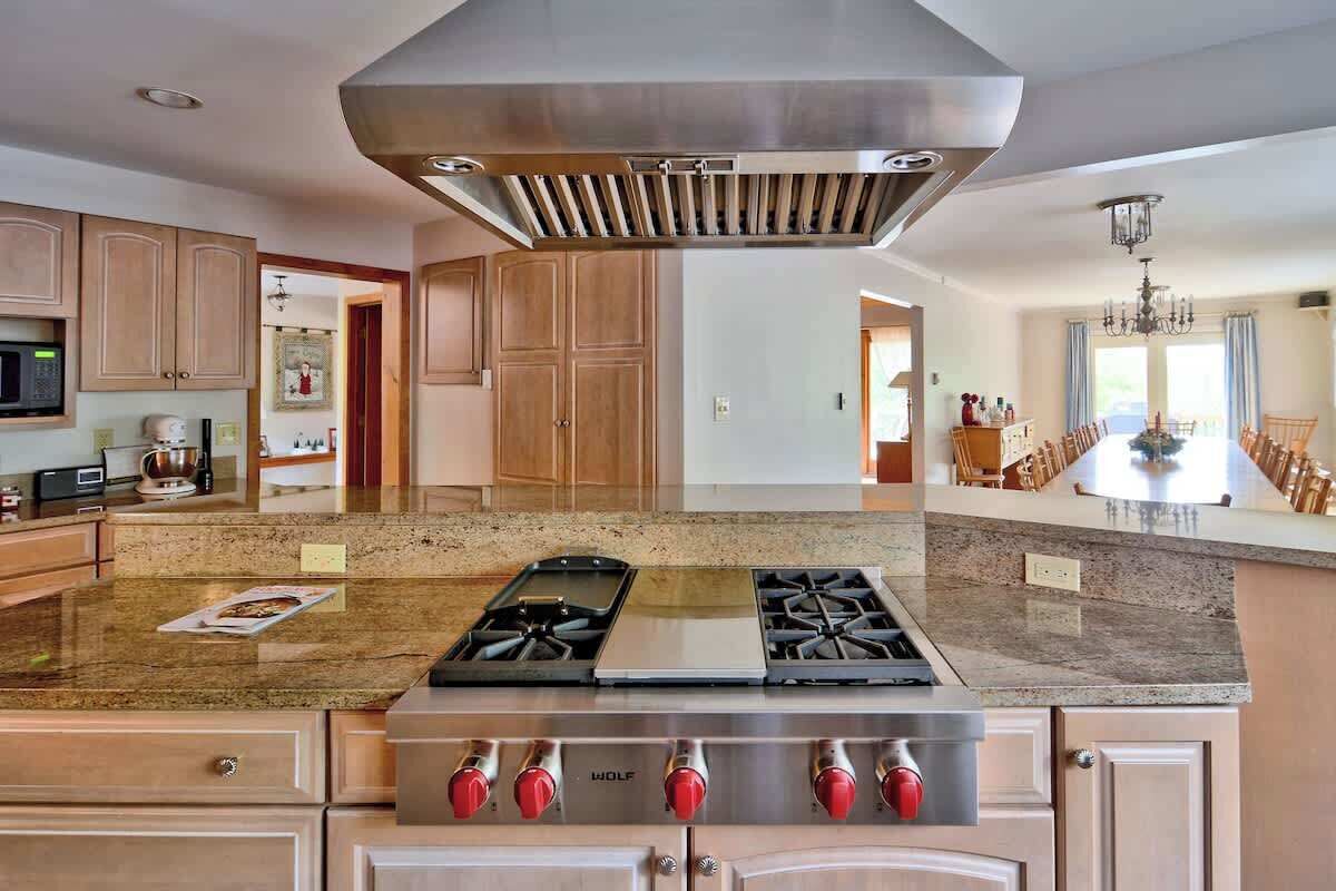 A kitchen with a gas stove, stainless steel range hood, wooden cabinets, granite countertops, and a dining area with a chandelier.