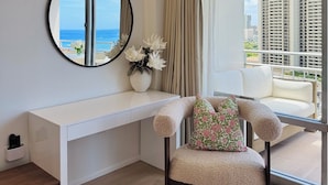 Une chambre moderne avec un bureau blanc, un miroir rond, une chaise agrémentée de coussins à motifs floraux et une grande fenêtre offrant une vue sur la ville et l’océan.