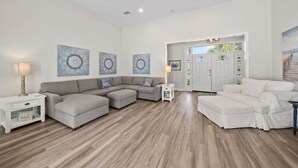 A spacious living room with a grey sectional sofa, a white armchair, and a white sofa. There are decorative wall art pieces and a wooden floor.