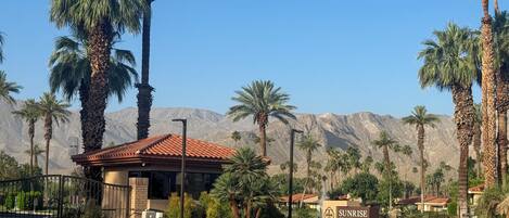 A building with a red-tiled roof, surrounded by palm trees and cacti, with a mountainous backdrop.
