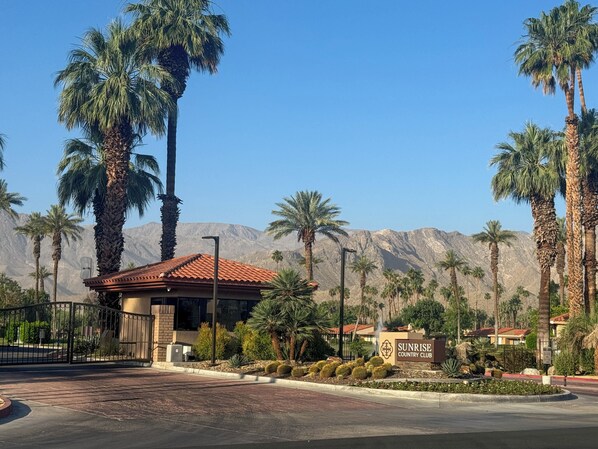 A building with a red-tiled roof, surrounded by palm trees and cacti, with a mountainous backdrop.
